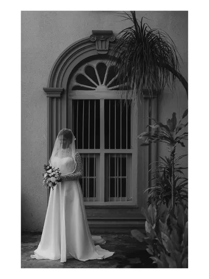 A timeless black and white bridal portrait. The bride, holding her bouquet, stands before an arched window, creating a classic and elegant composition.
