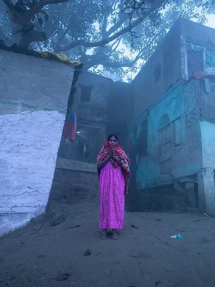 A woman in a bright pink saree stands alone in the dense fog on the Yamuna ghat, a solitary splash of color in a monochrome world.