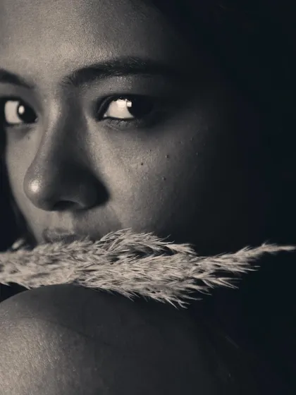 A black and white close-up using a piece of pampas grass to add texture and mystery. The lighting is soft and focused on the eyes.