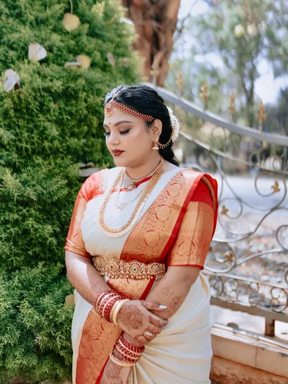 A serene portrait of a South Indian bride on her wedding day. The soft, glowing makeup and classic red lip perfectly match the elegance of her off-white and red Kanchipuram saree and traditional gold jewellery.