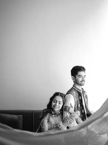 A soft and dreamy black and white portrait. The bride rests her head on the groom's shoulder, with a sheer curtain adding a gentle, romantic feel to the image.