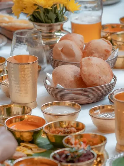 A close-up of a festive table set for a Sadya. The array of small bowls, each holding a unique preparation, shows the variety and complexity of the feast. Puffy pooris are ready to be paired with the delicious curries.