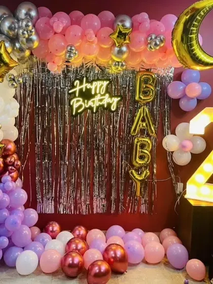 A fun and festive birthday decoration with a silver fringe curtain and a colorful balloon arch. The setup includes a light-up number '2' and 'BABY' foil balloons.