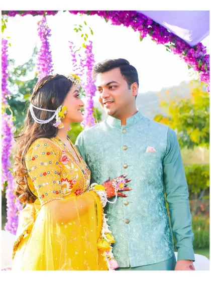 A joyful portrait of the couple at their Haldi ceremony. The bride is wearing a beautiful yellow lehenga with floral embroidery, complemented by the groom's pastel blue Nehru jacket.