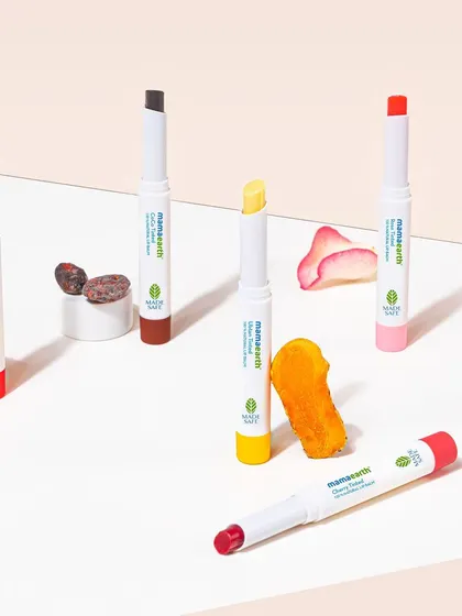 A group shot of Mamaearth's tinted lip balms, styled with their corresponding natural ingredients like orange, rose, and cocoa. The clean, geometric arrangement makes the product line easy to understand at a glance.