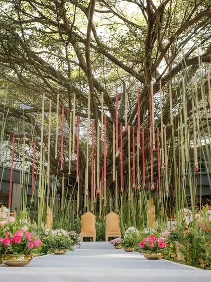 A serene and modern wedding aisle under a large tree, flanked by tall golden urns and leading to a stage with hanging floral strings.