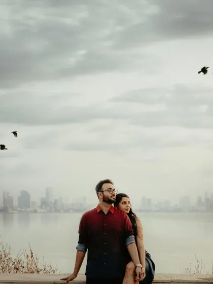A couple sits by the water in Mumbai, with the city skyline and birds in flight creating a serene urban backdrop. This photo showcases a relaxed pre-wedding shoot in a city environment.