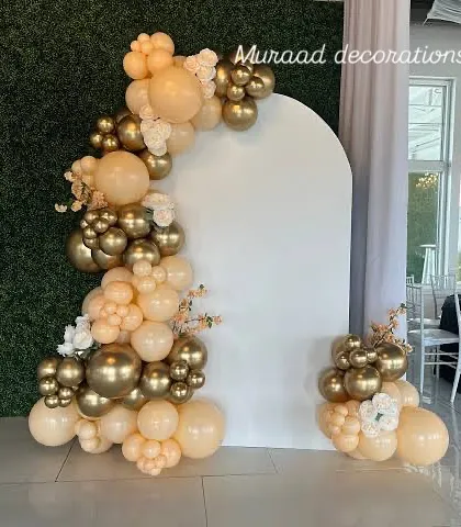 This modern backdrop features a simple white arched panel, asymmetrically decorated with a balloon garland in neutral tones of beige and chrome gold. A few white flowers add a touch of softness to this minimalist and chic setup.