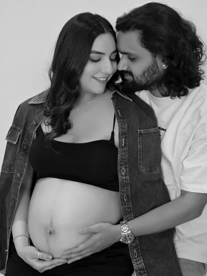 A close-up, intimate black and white shot. The focus is on their loving embrace and the hands cradling the baby bump.