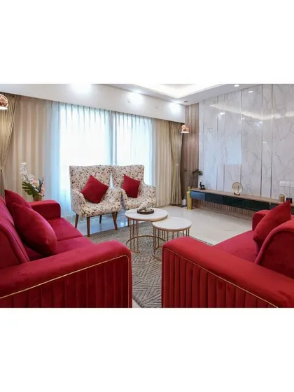 A living room designed for elegant hosting. The vibrant red velvet sofas with gold piping make a bold statement, contrasted with floral-patterned wingback chairs. The marble-clad TV wall adds a touch of modern luxury.