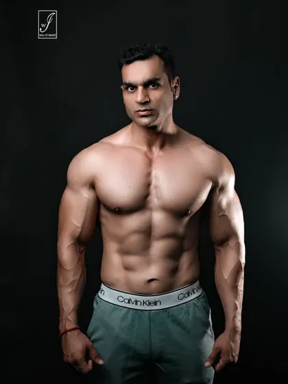 A powerful, direct-to-camera stance that showcases a full and well-developed chest and shoulders. The simple grey trousers and dark background keep the focus entirely on the upper body physique.