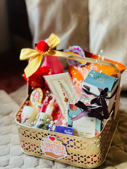 A traditional Rakhi hamper in a gold basket, filled with sweets, a rakhi, and a "brother like no other" sticker.