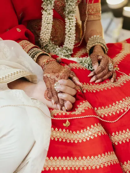 A close-up shot of the couple holding hands during the ceremony. The intricate Mehendi on the bride's hands and the rich red of her lehenga symbolize the beauty and tradition of the moment.