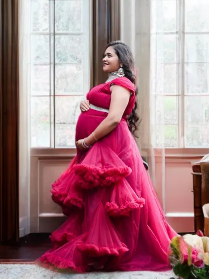 An indoor portrait by a window, featuring a mom-to-be in a gorgeous pink ruffled gown. The natural light from the window creates a soft and flattering glow.