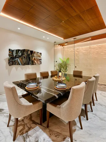 A perspective shot of a formal dining area. The square black marble table, seating eight, is positioned under a coffered wood ceiling with perimeter lighting, creating an intimate yet grand setting for meals.