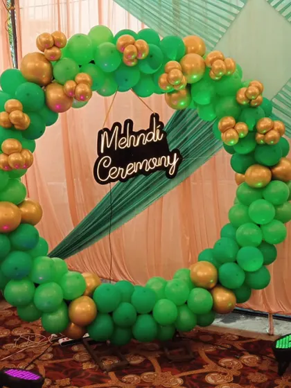 A close-up of a Mehndi ceremony backdrop I designed. This features a full ring of green and gold balloons, which are classic Mehndi colors, with a custom 'Mehndi Ceremony' sign in the center. It’s a perfect photo booth spot.