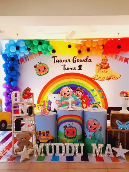 A bright and sunny Cocomelon party for Taanvi Gowda's first birthday. The setup includes a large rainbow, the yellow school bus, and a personalized backdrop.