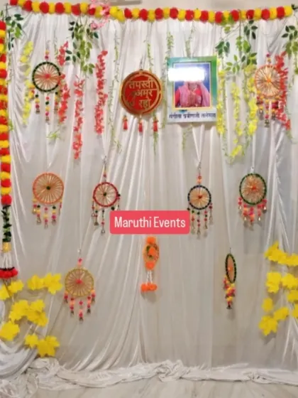 A custom backdrop for a Tapasvi homecoming, featuring a white satin drape decorated with handmade dream catchers, paper hangings, and floral vines. A photo of a guru is included as requested by the family.