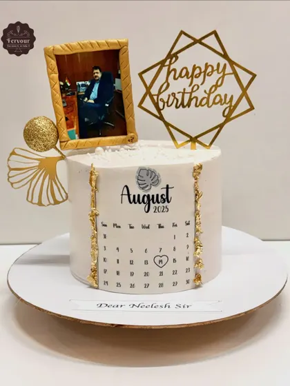 This unique birthday cake features a personalized calendar marking a special date, alongside a framed edible photo. The elegant white and gold design is perfect for a sophisticated corporate or personal celebration.
