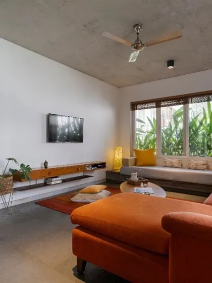 In this living room, a large window with a built-in bay seat is framed by lush greenery instead of traditional curtains. The vibrant orange sectional sofa adds a bold splash of color against the neutral palette of white walls and a concrete ceiling.