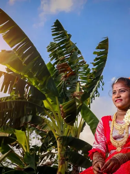 A wide shot capturing the subject in her traditional attire next to a banana tree. The bright sun and blue sky create a vibrant, tropical setting for this puberty ceremony photoshoot.