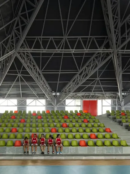 The tiered seating inside the 'Figured Ground' sports arena, with vibrant green and orange seats creating a lively pattern against the concrete steps. The exposed steel truss roof structure spans the entire hall, allowing for an unobstructed view of the court.