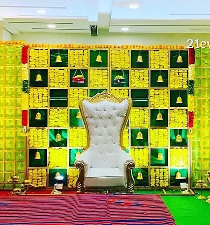 A bright and cheerful backdrop setup. The design uses a grid of yellow marigolds and green fabric, accented with hanging temple bells for a traditional touch.
