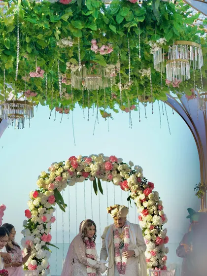 The couple taking their vows under a beautiful floral arch, part of a larger mandap structure with hanging crystal chandeliers and a canopy of green leaves.