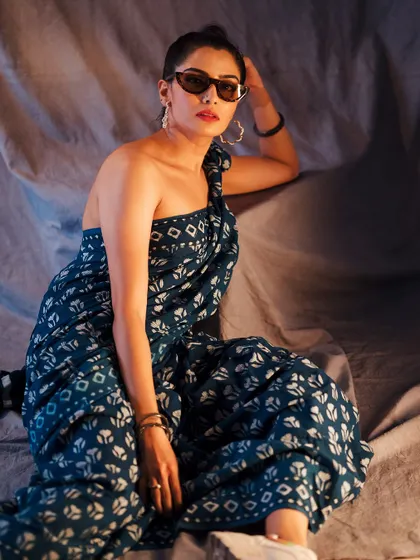 A full-length shot showing the modern drape of this printed saree. The relaxed, seated pose and sunglasses give it a contemporary, high-fashion feel.