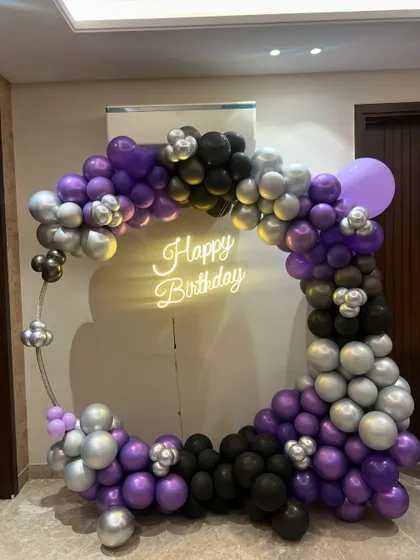 A stylish birthday photo booth with a circular frame and a garland of purple, black, and silver balloons, completed with a 'Happy Birthday' neon sign.