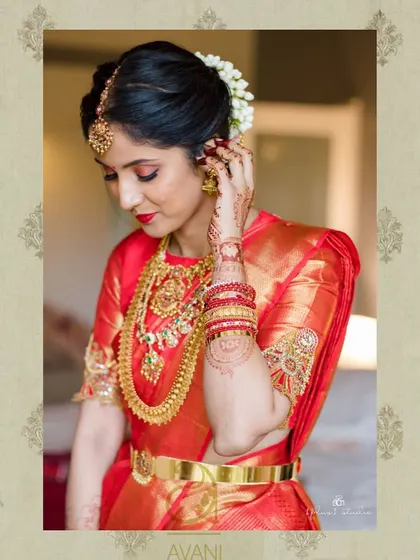 A bride getting ready, wearing a red Kanchipuram saree. Her custom blouse is adorned with heavy gold embroidery on the sleeves, perfect for her muhurtham.