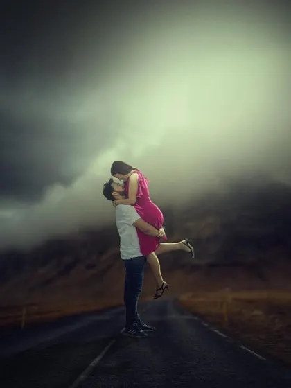 An epic, artistic pre-wedding photo of a groom lifting his bride on a deserted road under a dramatic, spotlight-like sky.