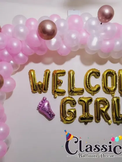 A 'Welcome Girl' decoration featuring a pink and rose gold balloon arch with matching foil letters.