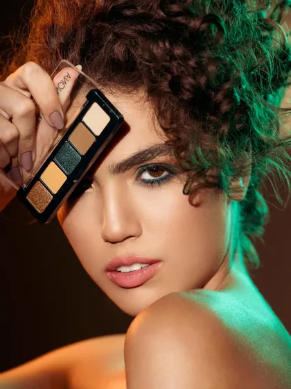 A striking portrait with dramatic lighting. The model holds an eyeshadow palette to her face, with a mix of warm and cool light creating a dynamic, high-fashion feel. Her confident gaze and the rich colors make this a captivating image for a makeup campaign.