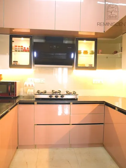 A beautiful U-shaped kitchen with glossy blush-pink lower cabinets and white upper cabinets. The design is both modern and warm, with under-cabinet lighting and black-framed glass display units.