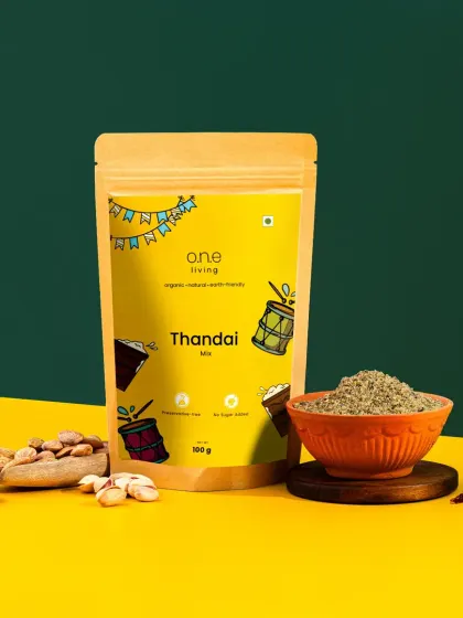 A clean hero shot of One Living's Thandai mix. The bold, color-blocked background and simple styling with raw ingredients put the focus entirely on the product's vibrant packaging.