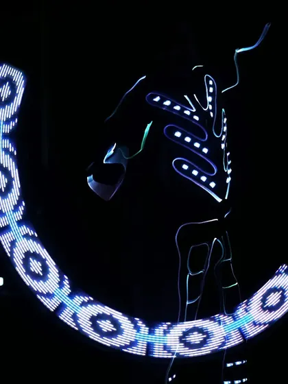 A Tron performer wields a visual poi, creating a sweeping arc of blue and white light patterns in the dark.