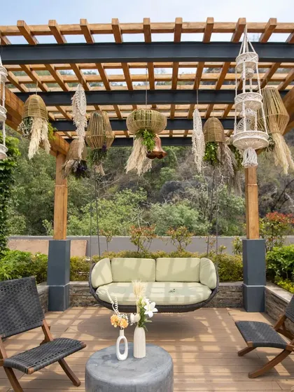 A cozy and stylish seating nook under a wooden pergola. The setup features a modern swing, comfortable chairs, and hanging macrame planters, creating a perfect spot to relax.