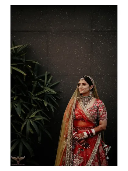 A simple, elegant portrait against a dark wall. The focus is entirely on the bride, allowing her vibrant red lehenga and her calm, happy expression to shine.