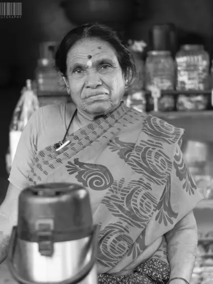 A black and white portrait of a female shopkeeper. The monochrome edit highlights the textures of her saree and the lines on her face, creating a powerful and dignified image.