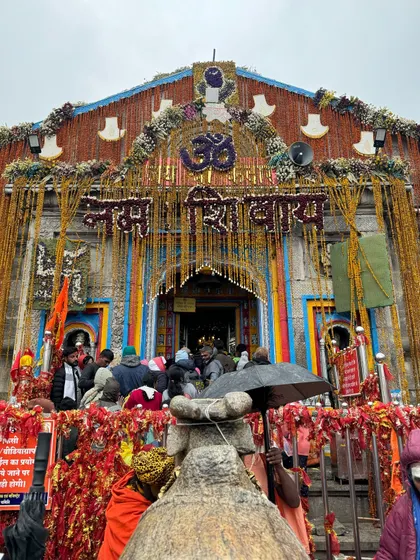The entrance to the Kedarnath temple, beautifully decorated with marigold flowers under a cloudy sky. The 'Om Namah Shivay' inscription welcomes devotees to the sanctum.