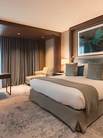 A luxurious and spacious master bedroom with a king-sized bed and a comfortable chaise lounge. The room features a neutral color palette, soft carpeting, and a large piece of abstract art.