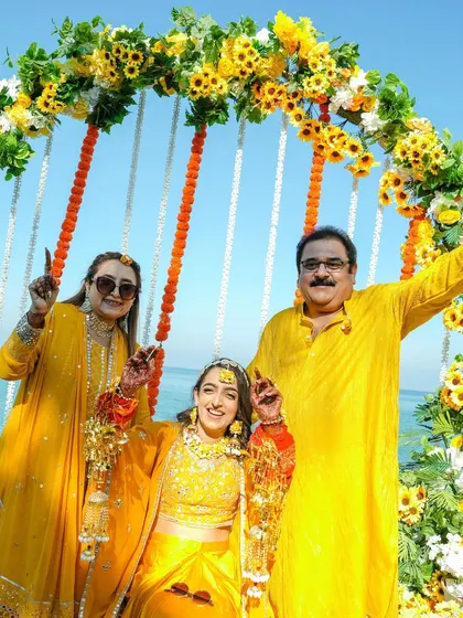 A happy family portrait from the Haldi ceremony, with the bride posing with her parents under a beautiful floral arch by the beach.