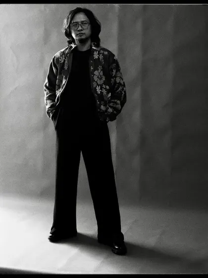 A full-length black and white portrait of Taba Chake. The simple studio background allows his quiet confidence to take center stage.