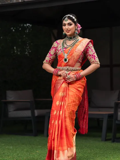 An outdoor bridal portrait where the vibrant orange and pink saree stands out against the natural green background. This shot showcases a complete bridal look, from the diamond matha patti to the embroidered blouse.