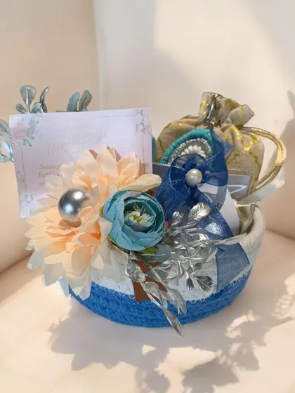 A small, charming hamper in a blue and white rope basket. This compact gift is decorated with flowers and contains small potlis, ideal for return favors or personal gifting.