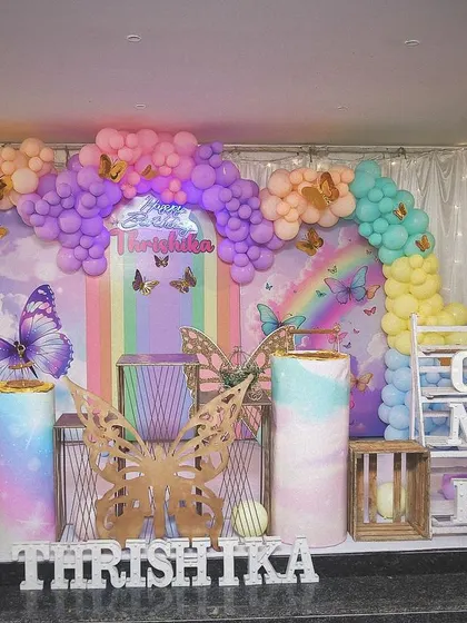 A full view of a rainbow butterfly theme, this setup includes custom-printed pedestals, a large balloon arch in various colors, and a personalized milestone board for the first birthday.