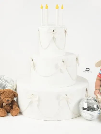 A simple and classic first birthday setup with a three-tiered prop cake, disco balls, and a teddy bear. The focus is all on the sweet birthday boy.