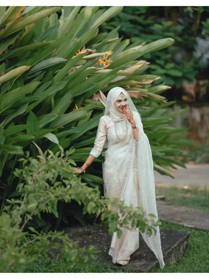 A serene portrait of a Muslim bride in a beautiful white outfit. The lush green background and her gentle, happy expression create a feeling of peace and timeless elegance.