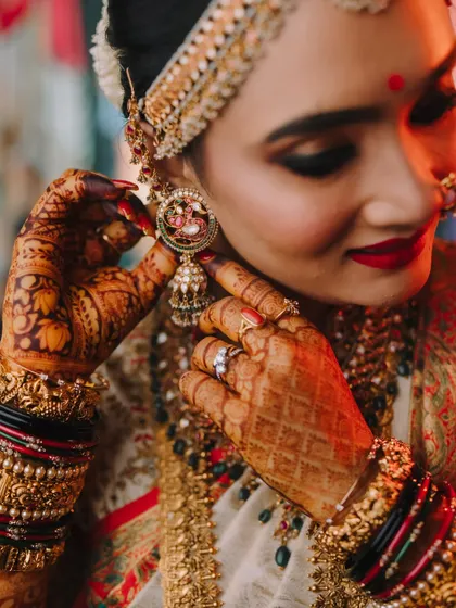 A detailed close-up of the bride's hands adjusting her earring, highlighting her intricate henna, bangles, and the craftsmanship of her jewelry.